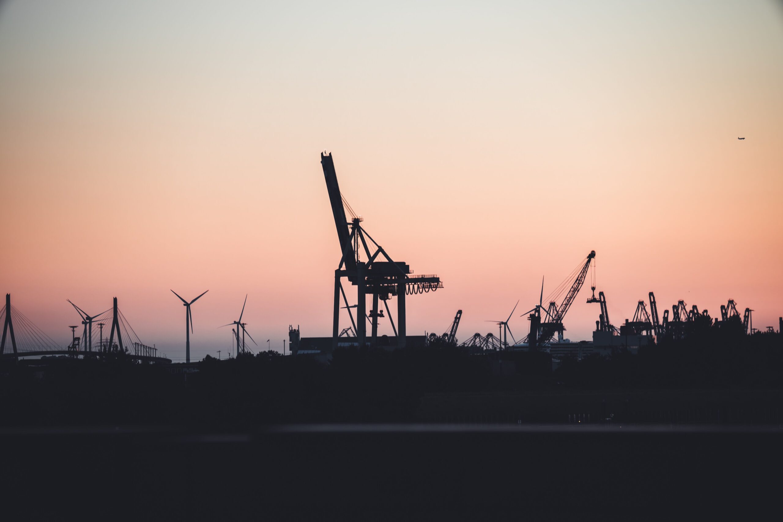 Silhouetted cranes and wind turbines at the port of Hamburg during sunset, highlighting industrial beauty.